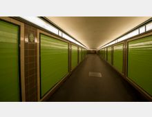 Unterführung mit grün gestrichenen Wandpaneelen auf beiden Seiten, schmalen Fliesenleisten und einer durchgehenden, indirekten Beleuchtung entlang der Decke. Der Boden ist aus dunklem Asphalt, und die Perspektive zeigt einen sich verjüngenden Gang in die Tiefe.