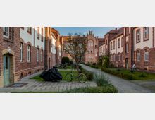 Hof eines Wohnkomplexes mit Backsteinfassaden, symmetrisch angeordneten Gebäuden und weißen Fenstern. In der Mitte befinden sich ein gepflasterter Weg, ein Baum, Fahrräder und kleine begrünte Flächen.