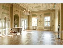 Elegantes historisches Zimmer mit reich verzierten Wänden und Decken in Pastelltönen, einem dekorativen Kamin, einem Flügel, Stuckarbeiten, Wandmalereien und Kronleuchtern. Große Fenster lassen viel Tageslicht herein, und der Boden ist mit einem Parkettmuster gestaltet.