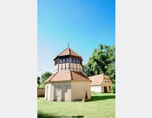 Ein kleines, achteckiges Fachwerkgebäude mit einem roten Ziegeldach steht auf einer Wiese, umgeben von Bäumen und Nebengebäuden mit ähnlicher Architektur. Der Himmel ist klar und blau, und die Szenerie wirkt ländlich.