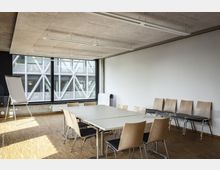 Moderner Besprechungsraum mit hellem Holzboden, rechteckigen Tischen und Holzstühlen. Große Fensterfront mit Kreuzmuster und Blick auf eine metallische Außenfassade; ein Flipchart steht links im Raum.