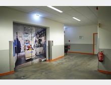 Flur in einem Gebäude mit grauem Betonboden und hellen Wänden, an denen Feuerlöscher angebracht sind. Eine große Tür zeigt ein großformatiges Bild des Innenraums eines Krankenwagens.