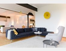 Wohnzimmer mit moderner Einrichtung: eine große, dunkelblaue Eckcouch mit Kissen, ein kleiner weißer Couchtisch und ein grauer Sessel. Im Hintergrund sind ein Essbereich mit Holztisch und Stühlen sowie eine offene Küche sichtbar; weiße Wände und eine gelbe Wanddekoration an der rechten Seite runden das helle, offene Design ab.