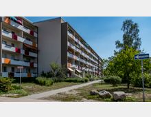 Mehrstöckiges Wohnhaus mit bunter Fassadengestaltung und Balkonen, umgeben von einer Grünfläche mit Bäumen und Sträuchern. Im Vordergrund ein Gehweg und ein Straßenschild mit der Aufschrift „Einbahnstraße“.