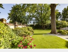 Ein gepflegter Garten mit einer großen Grünfläche, roten Blumen und Sträuchern, umgeben von hohen Bäumen. Im Hintergrund ist eine helle Steinmauer mit kleinen Ziegelaufsätzen zu sehen.