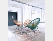 Ein moderner Innenraum mit hohen Decken, weißen Wänden und großen Rundbogenfenstern. Im Vordergrund stehen drei bunte Stühle im Retro-Stil um einen kleinen Glastisch auf einem gemusterten Teppich.