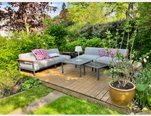 Holzterrasse in einem Garten mit zwei grauen Sofas, einem kleinen Tisch mit einer weißen Lampe und einem großen Blumentopf. Umgeben von üppigem Grün, Sträuchern und vereinzelten Blumen.
