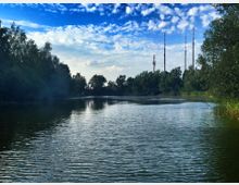 Ein ruhiger See umgeben von Bäumen, mit mehreren hohen Sendemasten im Hintergrund unter einem teils bewölkten blauen Himmel.