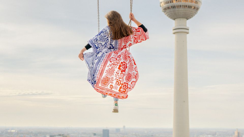 Blick über die Berliner Innenstadt mit dem Fernsehturm am Alexanderplatz im Hintergrund. Im Vordergrund hängt eine Schaukel an Ketten hoch über den Dächern, darunter sind Straßen, Gebäude und Baumreihen zu sehen.