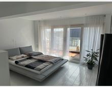 Helles Schlafzimmer mit einem niedrigen Bett und grauer Bettwäsche, bodentiefen Fenstern mit weißen Vorhängen und einem Zugang zu einer Terrasse. Eine Zimmerpflanze steht rechts im Raum auf einem hellen Holzfußboden.