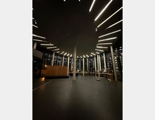 Großer, moderner Innenraum mit hohen Glaswänden und einer Decke, die von radial angeordneten, leuchtenden LED-Streifen betont wird. Im Vordergrund ein leerer, dunkler Boden, mit vereinzelten Säulen und Holzmodulen im Hintergrund.