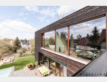 Modernes Wohnhaus mit dunkler Holzfassade und großen Glasfronten, die den Blick in ein hell eingerichtetes Zimmer freigeben. Im Vordergrund eine Terrasse mit Holzdeck, Pflanzen und einem Tisch, umgeben von einem grünen Garten und weiteren Häusern in ländlicher Umgebung.