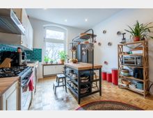 Helle, modern eingerichtete Küche mit Holzarbeitsflächen und einem freistehenden Küchenblock aus Metall. Die Wandfliesen sind türkis, große Fenster lassen Tageslicht herein, und Pflanzen sowie Küchenutensilien verleihen dem Raum eine wohnliche Atmosphäre.