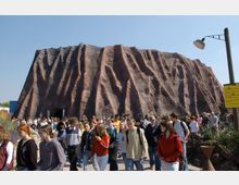 Eine große künstliche Felsformation mit welligen, rot-braunen Strukturen dient als Kulisse, während eine große Gruppe von Menschen, vorwiegend Jugendliche, über einen Weg davor läuft. Im Vordergrund sind Pflanzen und Lampen zu sehen, rechts ist ein gelbes Schild angebracht.