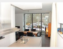 Offener Wohnraum mit moderner Küche, Essbereich und großen bodentiefen Fenstern, die in einen schneebedeckten Garten mit gestapeltem Brennholz blicken. Ein Esstisch mit schwarzen Stühlen und eine goldene Pendelleuchte sind zentrale Elemente im Raum.