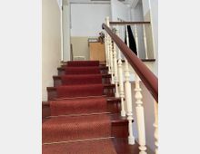 Eine Innentreppe mit dunkelrotem Teppichläufer, gesichert durch dünne Metallstangen. Das Geländer besteht aus hellen, verzierten Holzbalustern mit einem braunen Handlauf, und oben ist ein kleiner Flur mit einer geschlossenen Holztür sowie einem Schrank sichtbar.