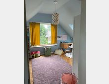 Ein Kinderzimmer mit hellblau gestrichenen Wänden, gelben Vorhängen vor einem großen Fenstersitz, einem lilafarbenen Teppich und Holzfußboden. Im Raum stehen ein Schreibtisch mit Stuhl, Spielzeugkisten und Sitzkissen, sowie eine bunte Hängelampe an der Decke.