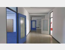 Ein leerer, heller Flur in einem Gebäude mit weißen Wänden und einem glänzenden Boden. Auf beiden Seiten befinden sich Türen mit blauen Rahmen und Glaseinsätzen, sowie eine geöffnete weiße Tür im Vordergrund.