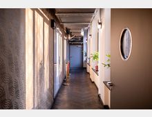 Ein schmaler, modern gestalteter Flur mit hellem Tageslicht, das durch Fenster auf einer Seite einfällt. Die rechte Wand ist mit Topfpflanzen dekoriert, während die linke Wand dezente wellenförmige Muster aufweist; eine graue Tür und sichtbare Deckeninstallationen befinden sich im Hintergrund.