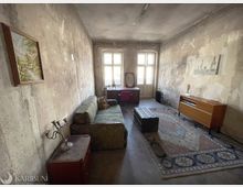 Langer, schmaler Wohnraum in einem alten Gebäude mit stark abgenutzten, fleckigen Putzwänden und zwei großen Fenstern sowie einer Glastür am Ende. Im Raum stehen ein Sofa, ein Schreibtisch mit Stuhl, eine Truhe als Couchtisch, ein großer Teppich und ein Sideboard.