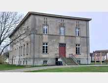 Zweistöckiges, rechteckiges Gebäude mit verputzter Fassade in beige-grauer Farbe, großen Fenstern und einer zentralen Eingangstür in Rot, die über eine kleine Treppe erreichbar ist. Im Vordergrund befindet sich eine Rasenfläche, im Hintergrund sind weitere Gebäude und ein Pavillon sichtbar.