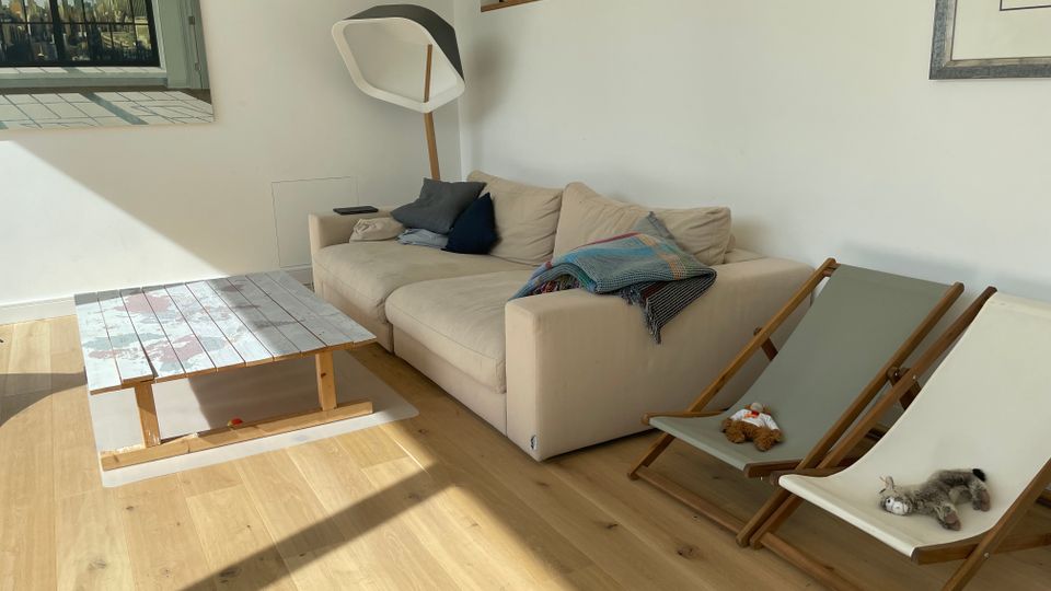 Helles, modern eingerichtetes Wohnzimmer mit hellem Holzboden, beigem Ecksofa und niedrigem Couchtisch aus Holz. An der Wand hängen gerahmte Bilder, daneben stehen zwei klappbare Liegestühle; eine Stehlampe und eine halbhohe Trennwand mit Ablage strukturieren den Raum.