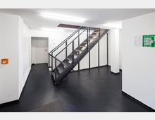 Treppenhaus in einem modernen Gebäude mit schwarzem Boden, weißen Wänden und einer Metalltreppe mit Gittergeländer. An den Wänden befinden sich Beschilderungen und ein Fluchtplan.