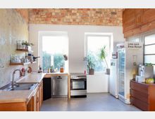 Wohnküche mit rustikalem Charme, bestehend aus gemauerten Wänden und sichtbaren Backsteinen. Zentrale Elemente sind zwei große Fenster, eine Spüle mit Arbeitsplatte, Herd, Kühlschrank und minimalistische Regale mit Küchenutensilien sowie Pflanzen als Dekoration.