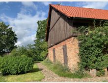 Ein Backsteingebäude mit roten Ziegeln und einem roten Satteldach, teilweise verkleidet mit Holz, umgeben von grünen Büschen und Bäumen. Ein gepflasterter Weg führt durch eine Wiese entlang der Vorderseite des Gebäudes.