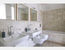 Modernes Badezimmer mit beigen Fliesen an Wänden und Boden. Zwei Waschbecken mit Spiegeln, ein WC, ein Bidet und eine Badewanne sind vorhanden.