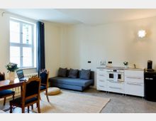 Heller Wohnraum mit offener Küchenzeile: links ein großes Fenster mit dunklem Vorhang sowie ein Esstisch mit Stühlen, mittig ein graues Sofa auf einem Teppich. Rechts eine weiße Küchenzeile mit Spüle und Backofen neben einem schwarzen Kühlschrank, darüber eine Wandleuchte.