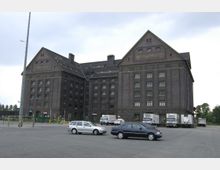 Backsteinlagerhaus mit der Aufschrift „Zollspeicher“ und „Westhafen“ auf den Giebelseiten, davor ein großer asphaltierter Platz mit geparkten Autos und Lkw, umgeben von Zäunen und Baumreihen im Hintergrund.