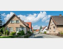 Straßenszene in einer Wohnsiedlung mit Einfamilienhäusern, die spitzgiebelige Dächer und bunte Fensterläden haben. Die Straße verläuft durch die Mitte, eingerahmt von Gehwegen, Vorgärten und Holzzäunen unter einem klaren Himmel mit Wolken.