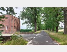 Roter Klinkerbau an einer Kreuzung, neben einer schmalen asphaltierten Allee, die von hohen Bäumen gesäumt ist; im Hintergrund sind Felder zu sehen.