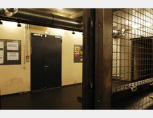 Ein industrieller Innenraum mit cremefarbenen Wänden, schwarzen Türen und Metallrohren an der Decke. Ein Gitter trennt den Vordergrund vom Hauptbereich, und an den Wänden befinden sich Hinweisschilder für Toiletten und die Garderobe.