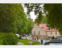 Ein Weg mit parkenden Autos führt durch eine grüne, baumreiche Umgebung zu einem beige-gelben Gebäude mit roten Ziegeldächern. Mehrere Personen, darunter Spaziergänger und Radfahrer, bewegen sich entlang des Weges.