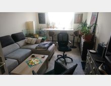 Ein kleines Wohnzimmer mit einer grauen Eckcouch, einem hellen Couchtisch und einem Bürostuhl in der Mitte. Pflanzen, ein Aquarium und eine Kommode mit Schubladen zieren den Raum, während ein Fenster im Hintergrund Tageslicht hereinlässt.