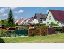 Reihenhaussiedlung mit Einfamilienhäusern, die teils farbig gestrichen sind, und roten oder dunklen Ziegeldächern. Im Vordergrund sind gepflegte Gärten mit Zäunen, Hecken und kleinen Gartenhäusern sowie eine Rasenfläche sichtbar, im Hintergrund eine Tanne und weitere Bepflanzung.