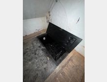 Kellerraum mit weiß verputzten Wänden und Steinboden; im Boden befindet sich eine rechteckige, geöffnete Bodenluke mit schwarzer Klappe, die in einen dunklen, gefliesten Schacht führt. An der Wand verlaufen sichtbare Kabel und Leitungen.