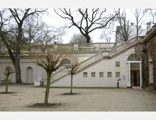 Hof einer weißen, historischen Gebäudefassade mit einer breiten Steintreppe und Balustrade, kahlen Bäumen im Vordergrund und einem offenen Torbogen auf der rechten Seite. Im Hintergrund sind lose angeordnete, hohe Bäume erkennbar.