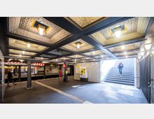 Unterirdische U-Bahn-Station mit moderner Deckenbeleuchtung und strukturierter Kassettendecke. Eine Treppe führt nach oben ins Freie, während ein Ticketautomat und Warnschilder auf dem Bahnsteig sichtbar sind.