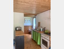 Schmale Küche mit Holzdecke und Holzboden, einer langen Arbeitsplatte rechts mit Herd und Spülbecken sowie einem grünen Vorhang als Unterschrankabdeckung. Am Ende des Raums stehen eine Holzverkleidung und eine Glastür nach außen, links befindet sich ein schmaler Tisch mit kleinem Gerät.