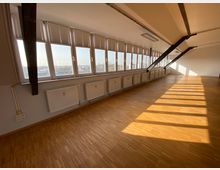 Ein leerer Raum mit schrägen Decken, bodentiefen Fenstern und Jalousien, die viel Tageslicht einlassen. Der Raum hat einen Holzfußboden, weiße Heizkörper entlang der Fensterfront und freiliegende Metallträger an der Decke.