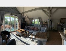 Wohnzimmer in einem Dachgeschoss mit schrägen Decken und sichtbaren Holzbalken. Der Raum enthält eine große hellgraue Couch mit dekorativen Kissen, einen Couchtisch aus Metall mit Glasplatte, einen schwarzen Sessel und einen Kronleuchter. Durch große Doppelfenster und Glastüren fällt Tageslicht ein, mit Blick auf eine Terrasse mit Sitzmöbeln und Grünpflanzen.