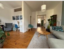 Wohnzimmer in einem modernen Stil mit Holzdielenboden und minimalistischer Möblierung. Zu den Elementen gehören eine graue Ecksofa-Ecke mit Kissen, ein Holzcouchtisch, Zimmerpflanzen, ein grauer Sessel, ein kleiner Schreibtisch sowie LED-Pendelleuchten und Wanddekoration. Im Hintergrund führt eine offene Tür in einen Essbereich mit großem Fenster.