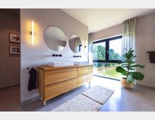 Modernes Badezimmer mit einer Holz-Waschtischkonsole, zwei runden Spiegeln und rechteckigen Waschbecken. Große Fenster mit Blick ins Grüne, lichtdurchfluteter Raum, ergänzt durch eine Zimmerpflanze in einem geflochtenen Korb und helle Vorhänge.