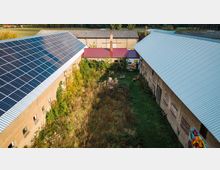 Eine ländliche Hofanlage mit zwei gegenüberliegenden Gebäuden, deren Dächer mit Wellblech gedeckt sind, eines davon mit Solarpaneelen. Der Innenhof ist überwuchert mit hohem Gras und Sträuchern, während die Gebäude teils mit Graffiti bemalt sind.