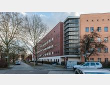 Straßenszene mit mehrstöckigen Gebäuden im Bauhaus-Stil, darunter ein rotes Backsteingebäude mit schmalen, horizontal ausgerichteten Fenstern und einem markanten Eckturm mit Glasblockfenstern. Im Vordergrund sind eine gepflasterte Straße, parkende Autos, einzelne Bäume ohne Laub und ein Gehweg zu sehen.