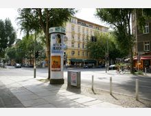 Straßenszene in einer urbanen Umgebung mit einem Werbesäule in der Mitte, die mit Plakaten beklebt ist. Im Hintergrund sind ein gelbes, mehrstöckiges Wohngebäude, Bäume, eine Kreuzung mit Verkehrsampeln sowie ein Straßencafé und geparkte Autos zu sehen.
