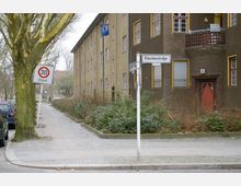 Straßenszene mit einem braunen Wohnhaus mit gelb umrandeten Fenstern und einem Balkon im Vordergrund. Ein Straßenschild mit der Aufschrift „Klemkestraße“ und ein Tempo-30-Verkehrsschild stehen an einer Kreuzung, daneben ein Gehweg mit parkenden Autos und einem großen Baum.